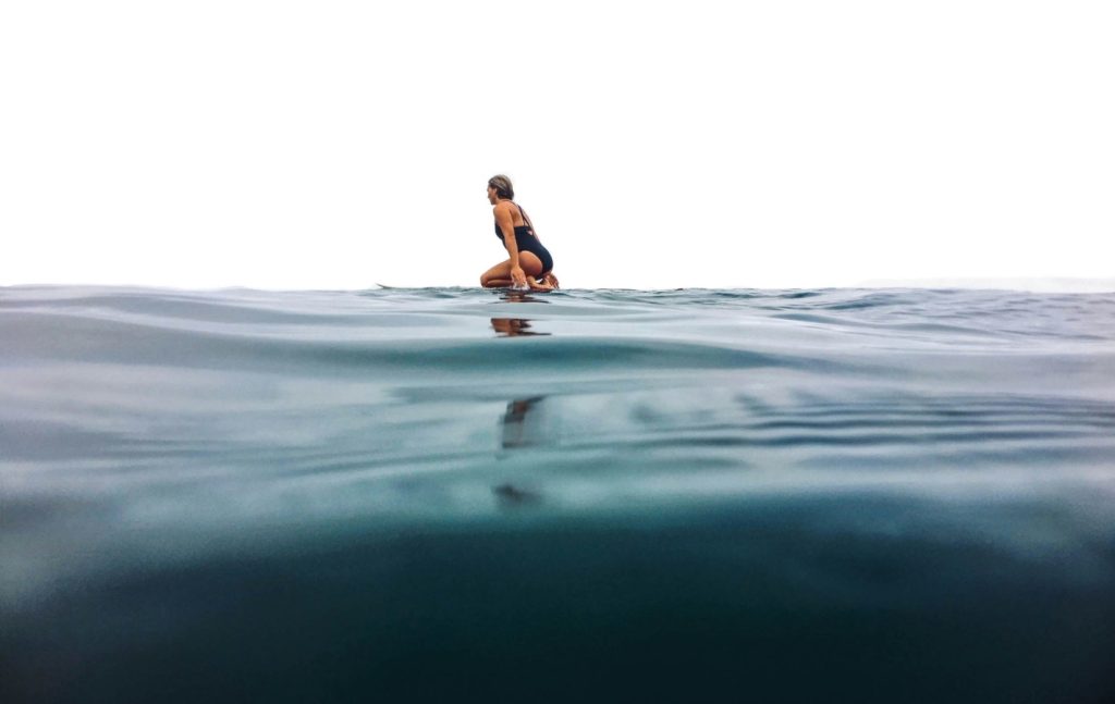A solo surfer kneels on her board, surrounded by calm sea and white sky — reflecting the quiet strength and inner focus at the heart of every outrider.